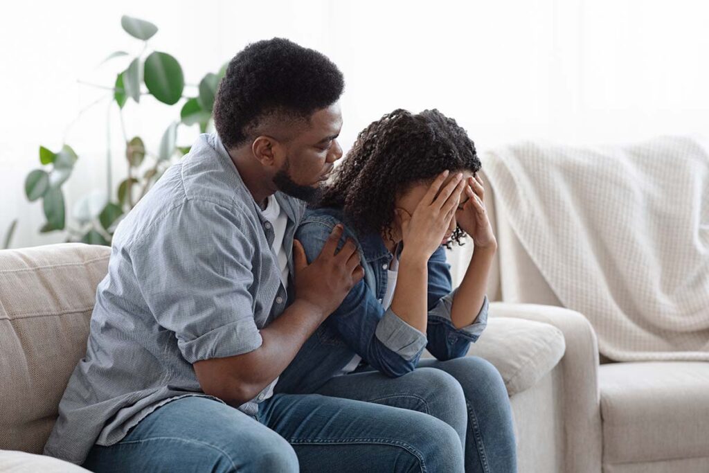 man seated next to his distraught wife on their couch strugglig to know how to help a spouse addicted to prescription drugs