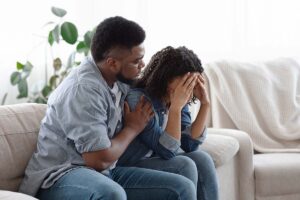 man seated next to his distraught wife on their couch strugglig to know how to help a spouse addicted to prescription drugs