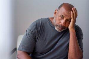 Man sitting with his head resting on his hand looks tired and pensive, wearing a dark gray shirt in a softly lit room.
