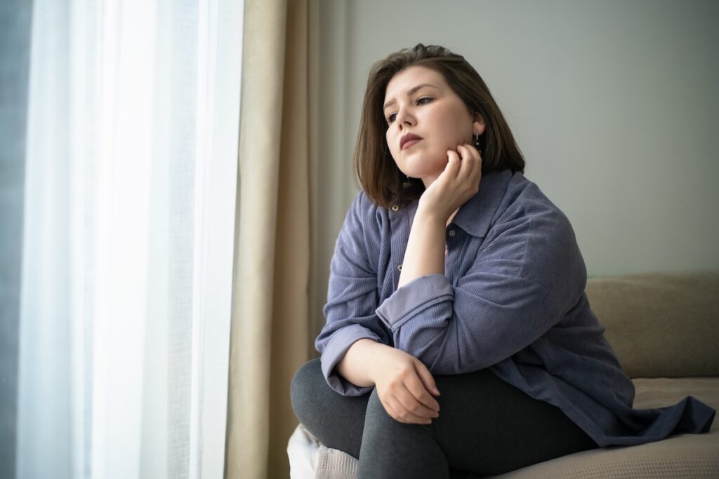 Woman sitting on a couch near a window rests her chin on her hand, looking thoughtful and pensive.