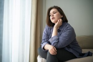 Woman sitting on a couch near a window rests her chin on her hand, looking thoughtful and pensive.